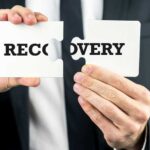 Image is a person in a black suit, holding a sign that is joining the words for "recovery"